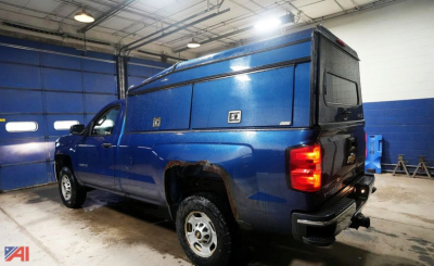 Blue 2016 Chevrolet Silverado 2500HD pickup with ARE cap and slider in an indoor shop, rear three-quarter view.