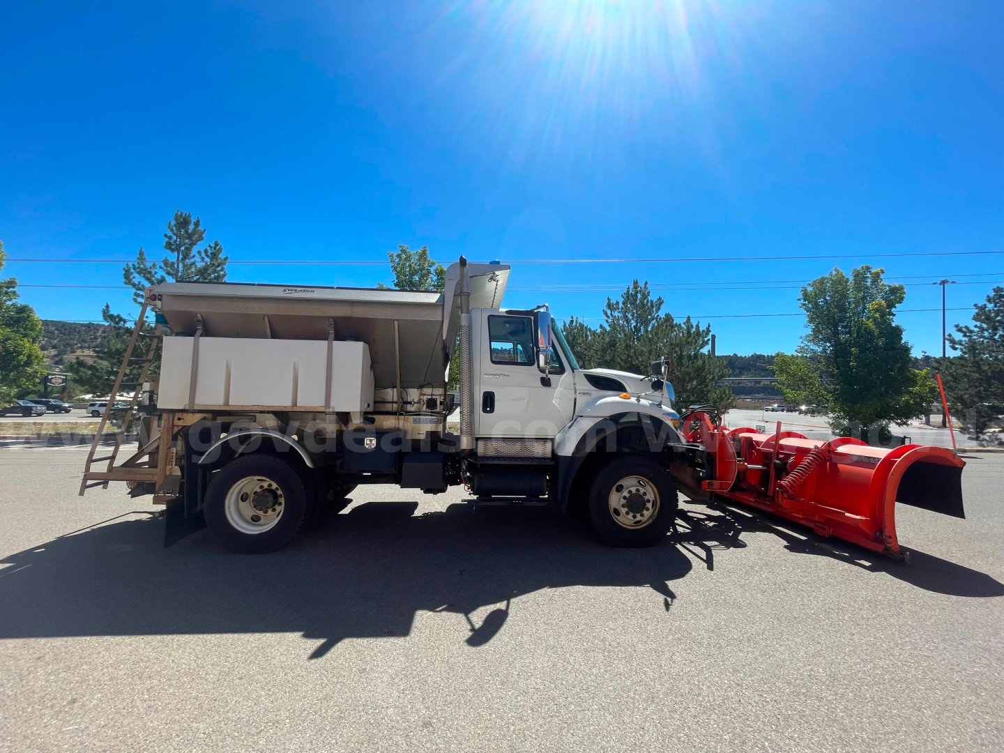 Truck in side view with a red snow plow attachment, dump bed, and International logo on cab. Clear blue sky, no visible text on equipment.