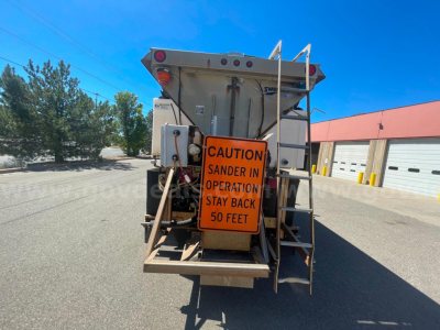 Dump truck rear showing orange caution sign reading 'CAUTION SANDER IN OPERATION STAY BACK 50 FEET' and metal framework.