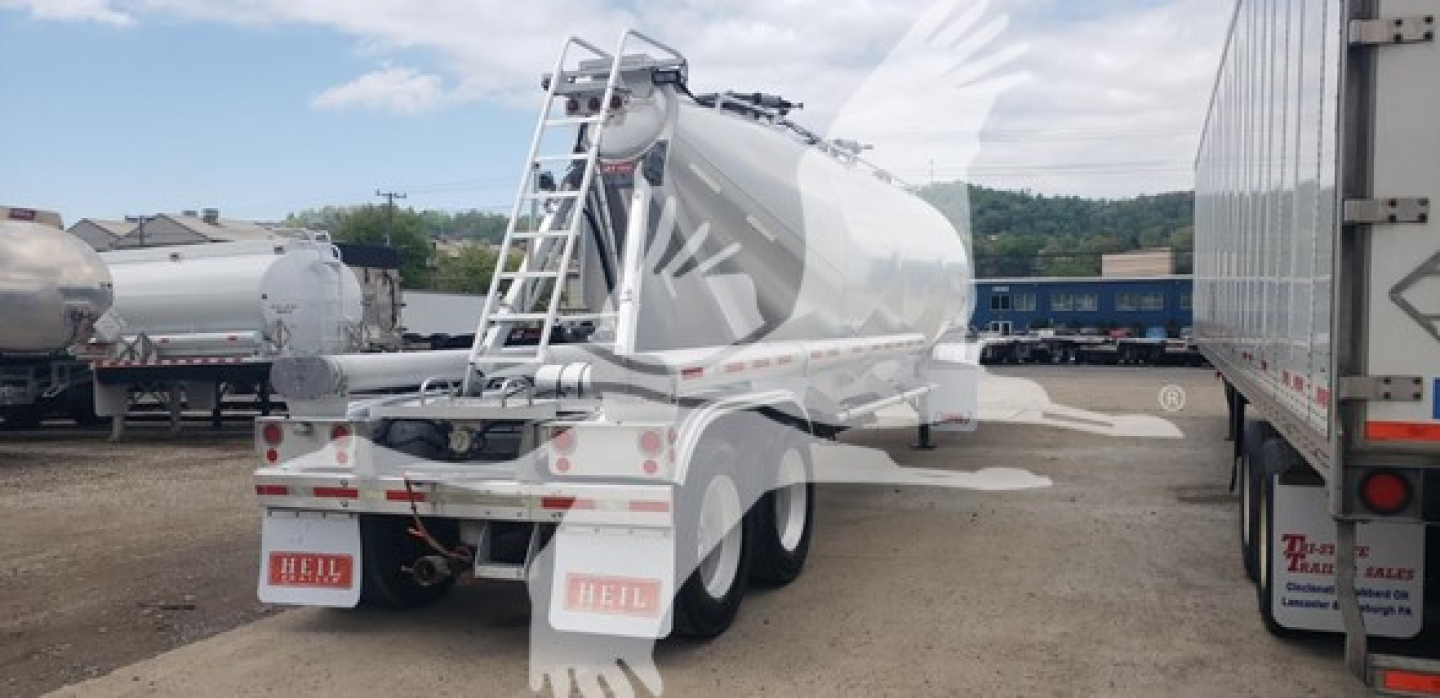 Pneumatic dry bulk trailer in a yard, rear and side view showing polished aluminum tank, ladders, and dual rear axles.