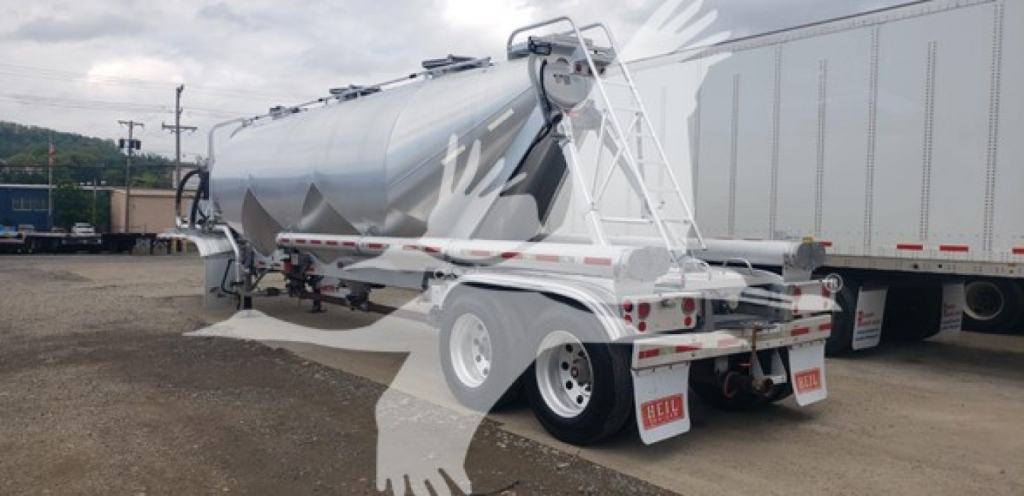 Pneumatic dry bulk trailer in transit with cylindrical tank and rear dual axle, white with safety rails and ladders, side hoses, and salvage yard background.