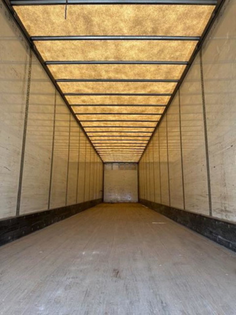 Interior of a long empty trailer with wooden ceiling panels and side walls, light entering from the far end, showing a spacious cargo area.
