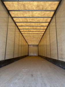 Interior of a long empty trailer with wooden ceiling panels and side walls, light entering from the far end, showing a spacious cargo area.