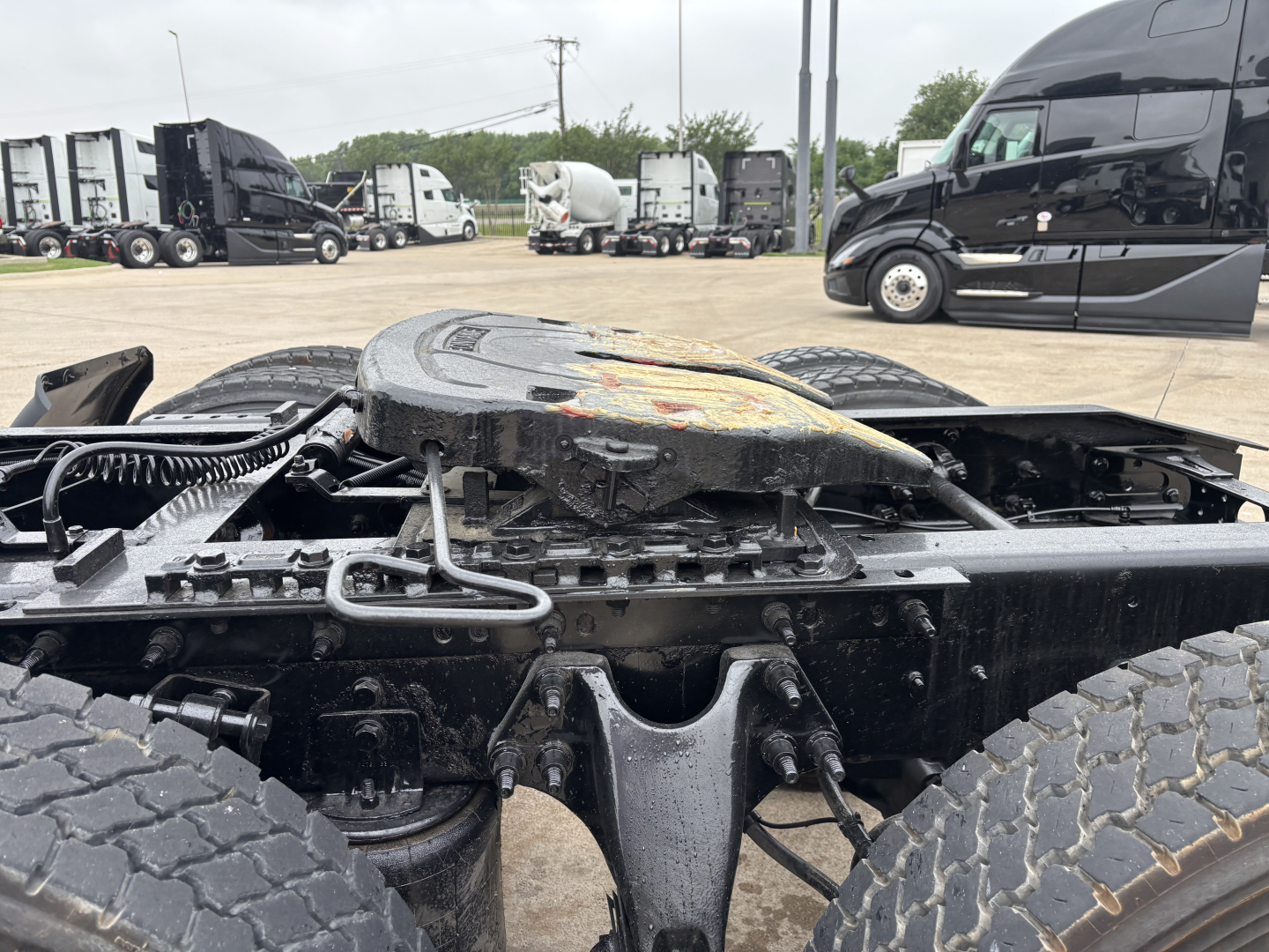 Truck chassis with fifth wheel and suspension visible; background shows multiple semi-trucks in a dealership lot.