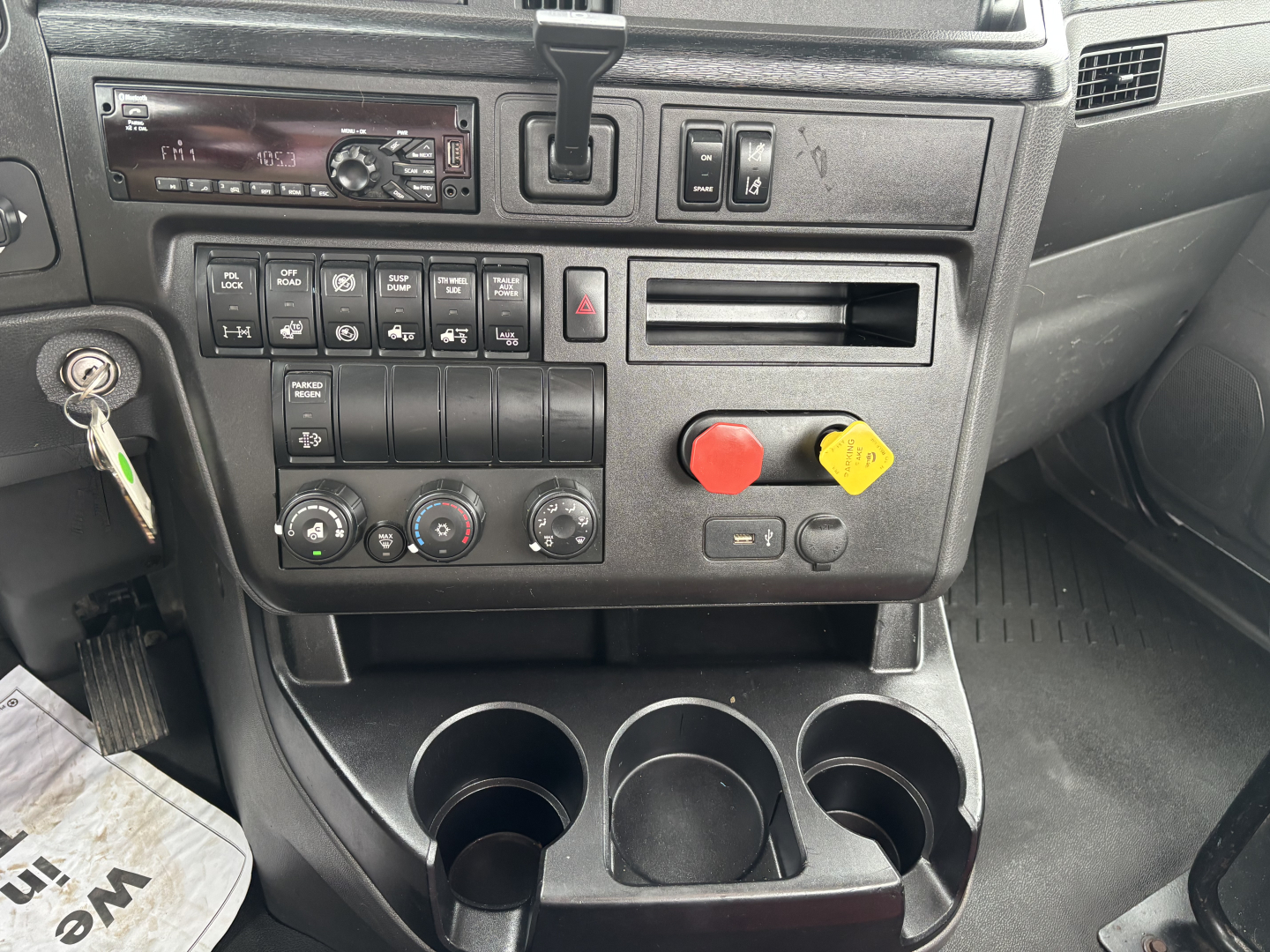 Truck cab dashboard with multiple switches and controls; gear selector; cup holders; keypad and red emergency stop knob; circular climate control dials; small display unit.