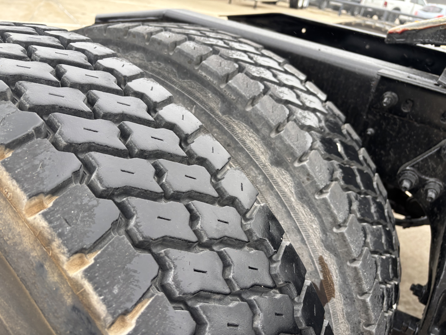 Truck wheel and tire close-up showing heavy tread on dual rear axle, with chassis frame and suspension components visible.