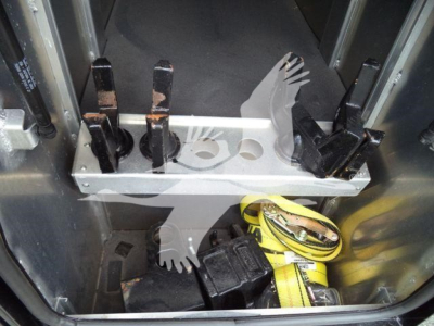 Front interior storage area of a Freightliner M2 106 winch truck showing metal tool rack with hooks, safety straps, and yellow ratchet strap.
