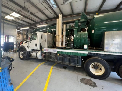 Truck side view of a vacuum tanker: white cab, green tank, with tall vertical exhaust stack and piping along side. Visible label on a small sign near the frame.