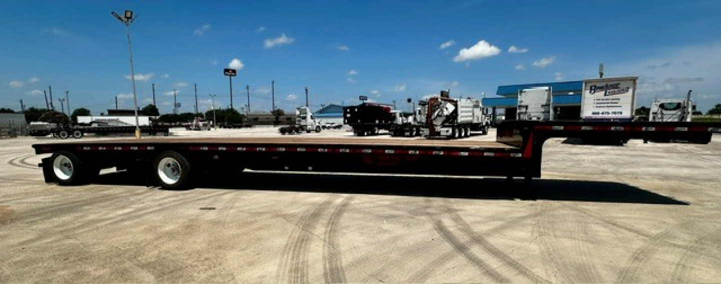 Flatbed trailer in side view showing long deck and dual rear axles with branding on site.