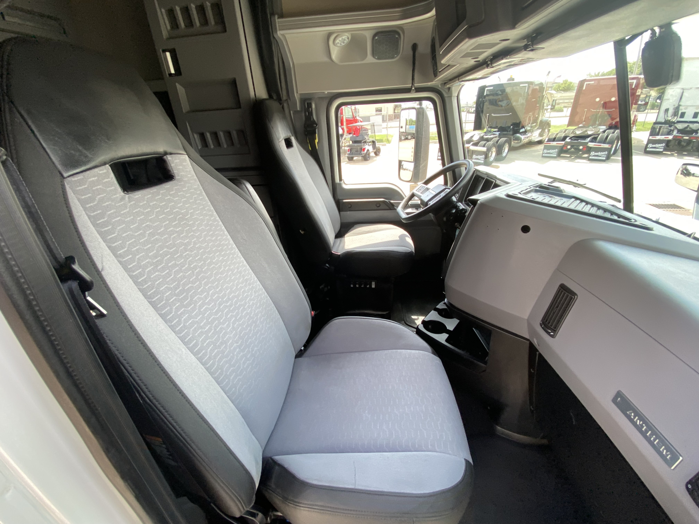 Truck cab interior of a 2020 Mack Anthem showing the driver seat and passenger seat with light gray upholstery, dashboard, and steering wheel visible.