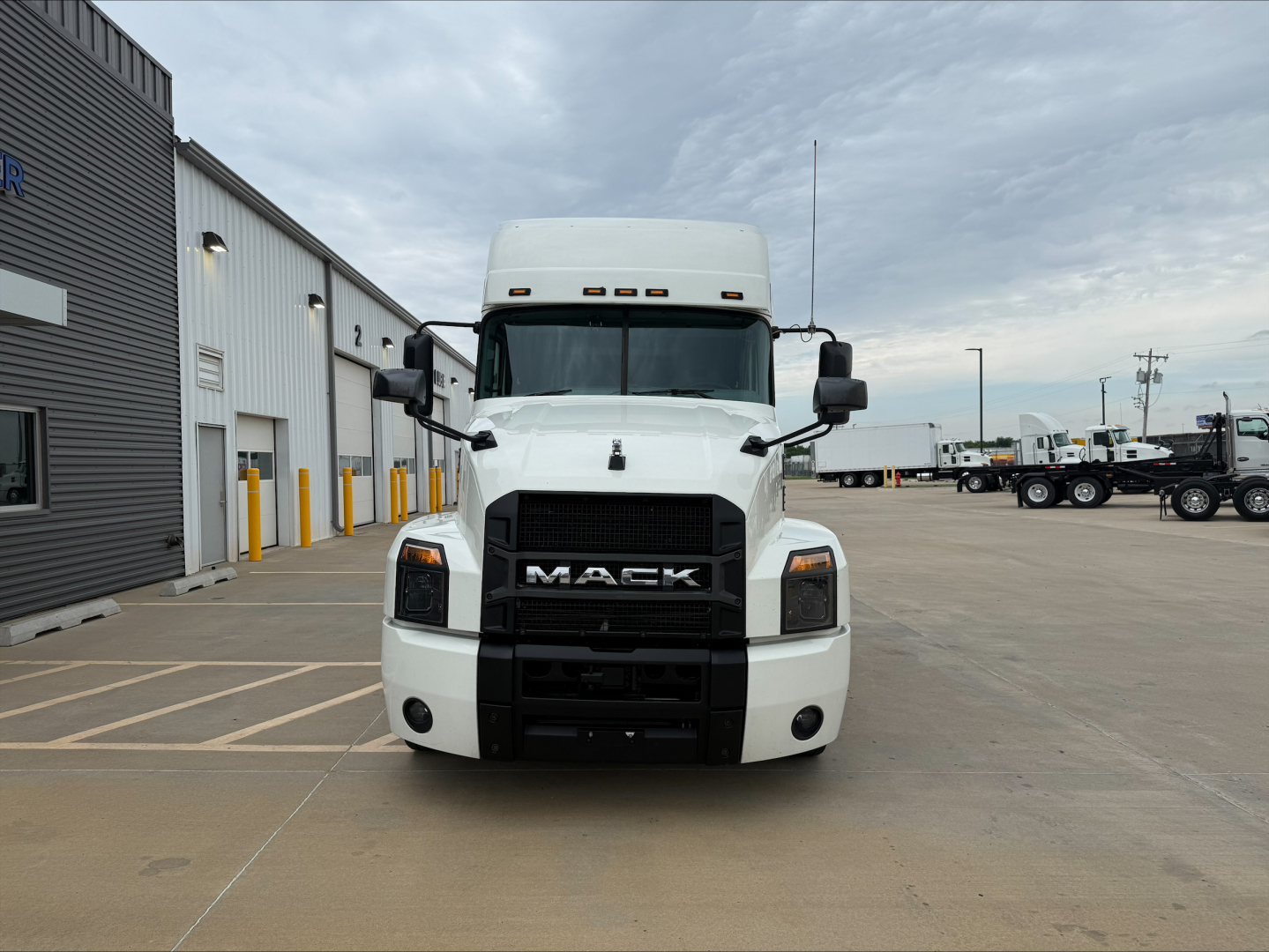 Front view of a white 2020 Mack Anthem conventional sleeper truck with black grille, Mack logo, and side mirrors; vehicles and dealership building in background.