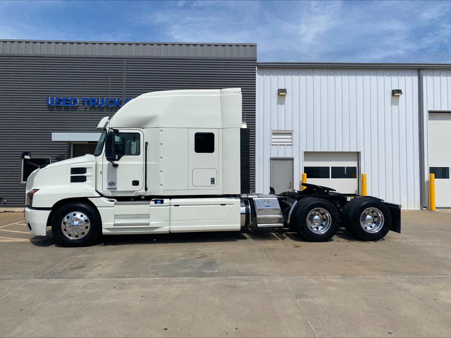 Truck cab and chassis of a 2020 Mack ANTHEM sleeper tractor, side view, with white paint and dual rear axles, parked in front of a dealership.