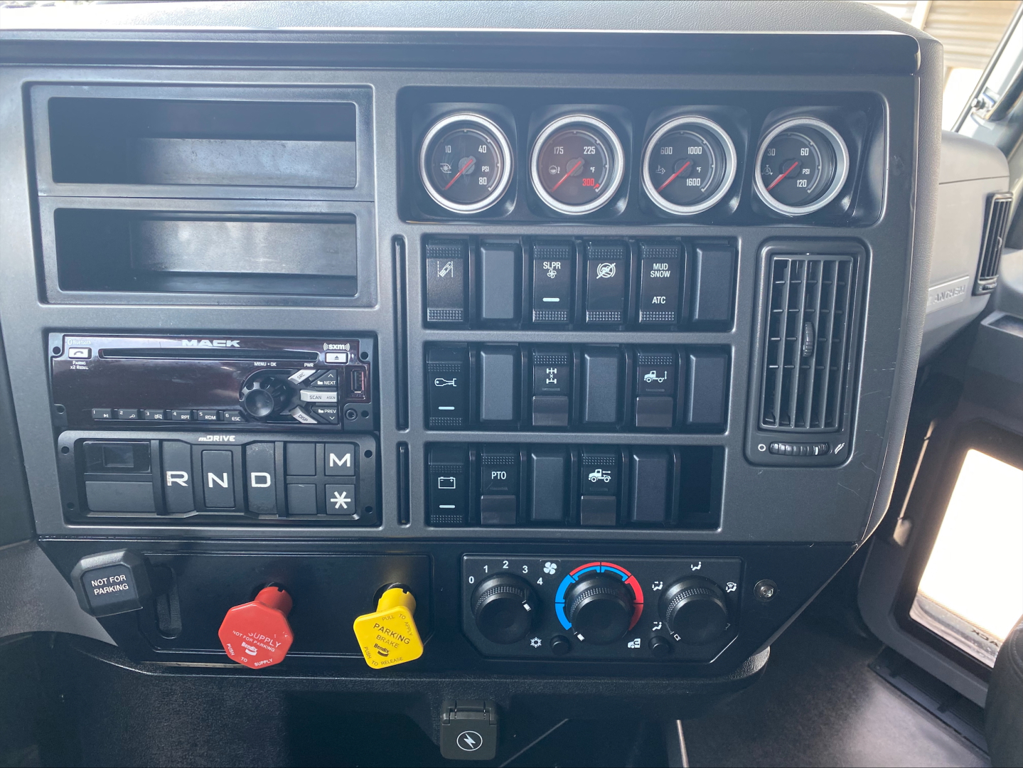 Dashboard of a Mack truck with several gauges, switches, and climate controls. Visible text includes R N D M on the gear selector, two colored knobs (red and yellow), and an auxiliary panel with various labels.