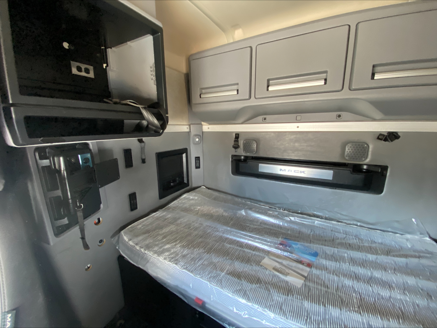 Truck cab interior showing sleeper bed and overhead cabinets in a Mack ANTHEM. Visible panels, storage compartments, and window shade.