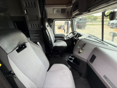 Truck cabin interior of a 2020 Mack Anthem, front seats and dashboard visible, steering wheel, driver and passenger seats, gray fabric upholstery, large windshield, side windows.