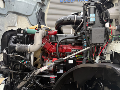 Engine bay of a 2020 Mack Anthem truck showing red engine with hoses, wiring, and components; visible label stickers and surrounding mechanical parts.