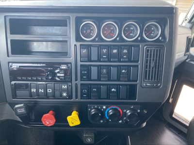 Dashboard of a Mack truck with several gauges, switches, and climate controls. Visible text includes R N D M on the gear selector, two colored knobs (red and yellow), and an auxiliary panel with various labels.
