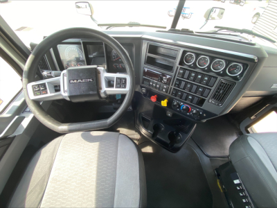Dashboard and steering wheel of a Mack truck interior showing a multi-gauge cluster, center console with switches, and grey seats.