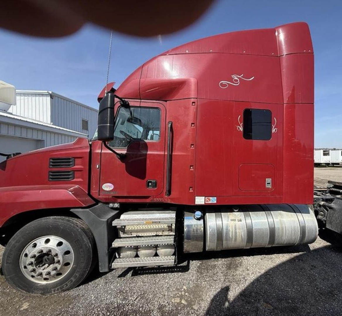 Side view of a red 2020 Mack Anthem sleeper cab tractor showing fuel tanks and steps. Visible text includes nothing legible besides generic truck features.