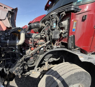 Truck engine bay with exposed components and hoses; red Mack Anthem cab visible on right; front wheel and suspension area; various labels and wiring in view.