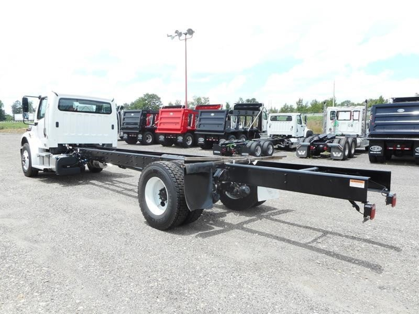 Freightliner M2 106 cab and chassis showing bare frame and front wheel with white cab, multiple trucks in background.