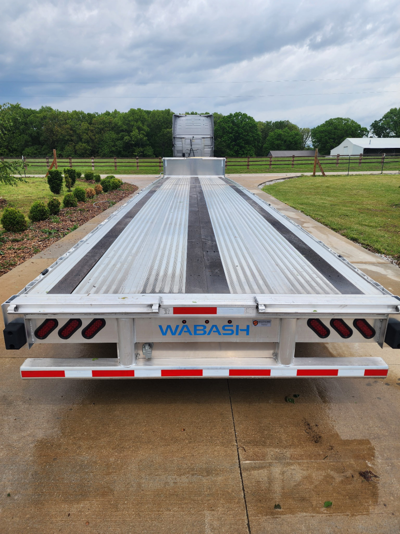 Flatbed trailer back view with WABASH logo, aluminum deck, side rails, tail lights, and ramp at end, long trailer in open lot