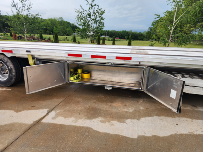 Wabash trailer body with open under-deck storage compartments showing yellow winch and some items inside; side rails and reflective tape visible.