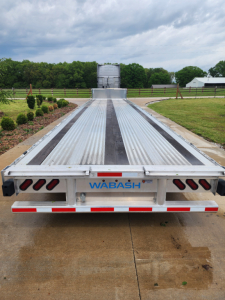 Flatbed trailer back view with WABASH logo, aluminum deck, side rails, tail lights, and ramp at end, long trailer in open lot