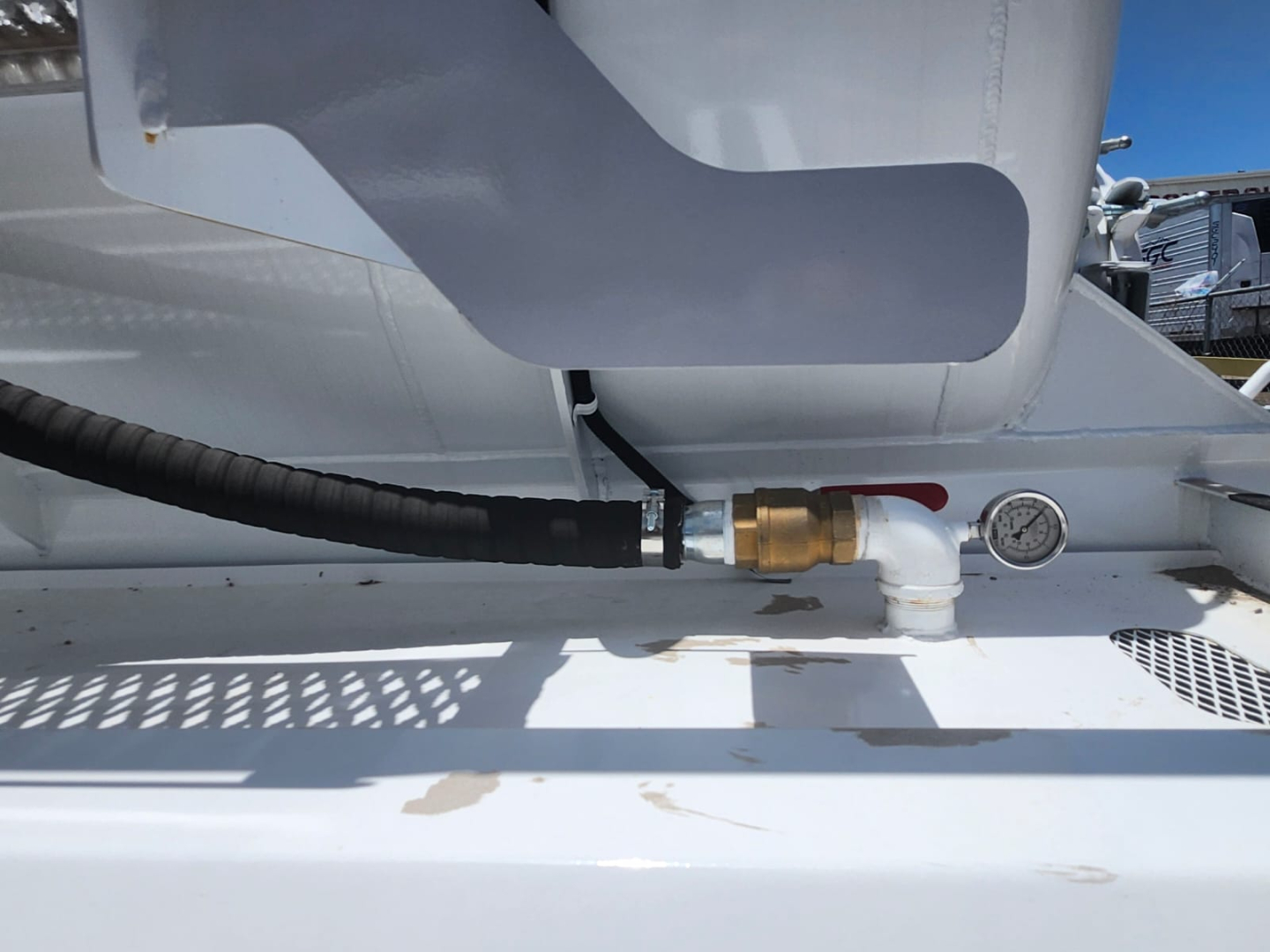 Tank fitting with brass valve and white pipe, hose and pressure gauge on a vacuum trailer side. Visible hoses and metal platform.