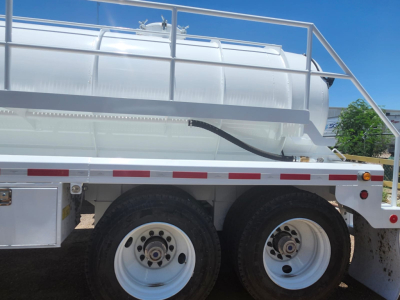 Vacuum trailer with large cylindrical stainless steel tank mounted on dual rear axles and white wheels, side rails, and rear platform.
