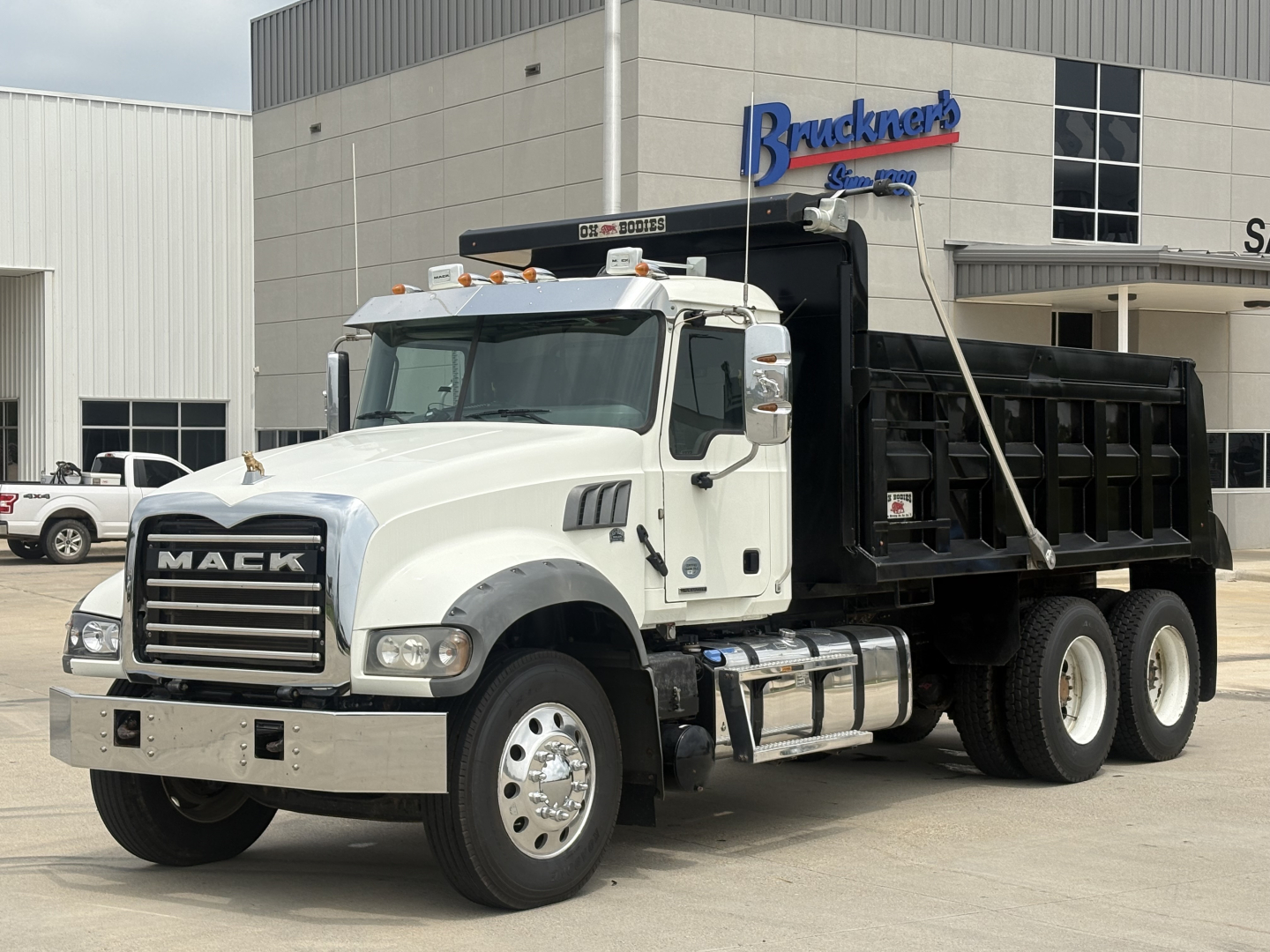 Mack GR64F dump truck in a dealership lot with white cab and black dump bed; visible text includes MACK on grille and Bruckner dealership building in background.