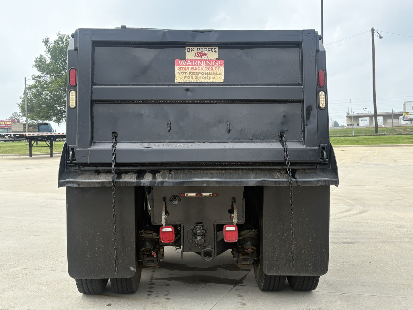 Mack GR64F dump truck rear showing dump bed tailgate and dual red warning reflectors, warning label on tailgate, and hitch/connector area between black mud flaps.