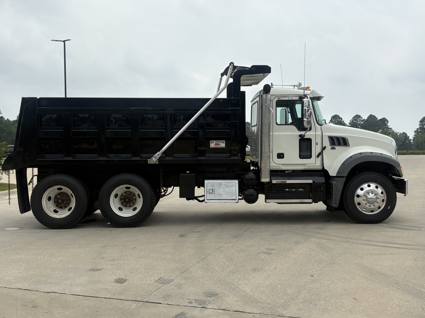 Dump truck side view showing 2020 Mack GR64F with dump bed, white cab, black dump body, chrome exhaust stack, dual rear axles, and fuel/tank area.