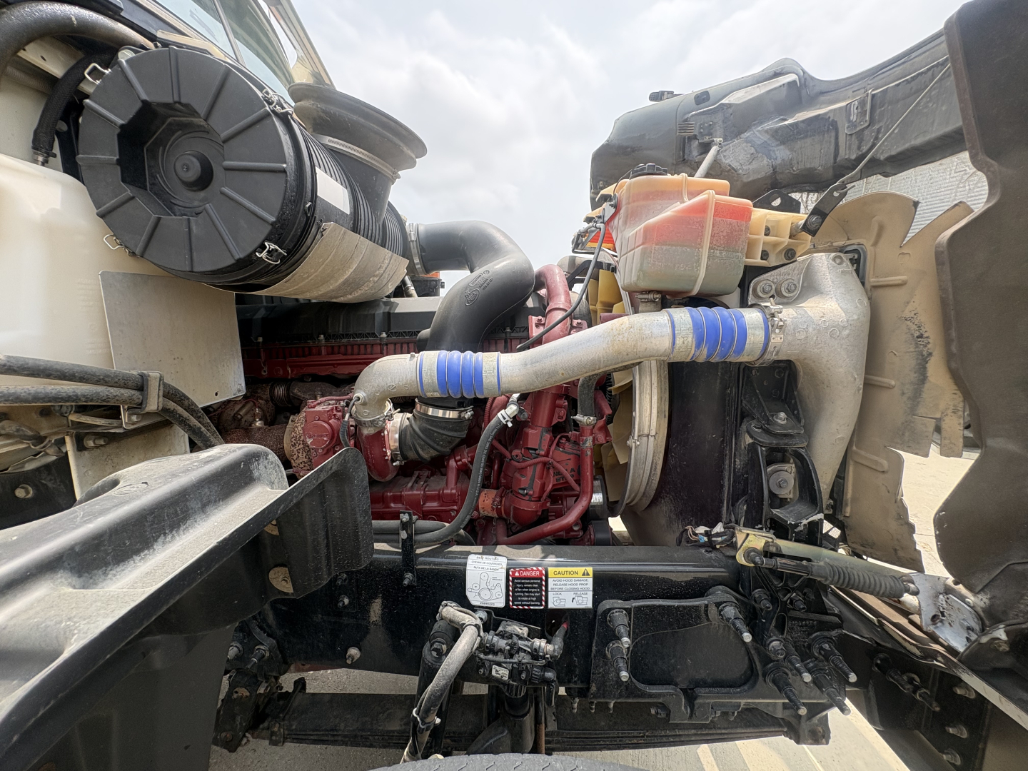 Mack GR64F engine bay showing coolant reservoir, hoses, and turbocharger. Visible components include radiator hose, coolant reservoir with orange cap, and engine block with red paint.