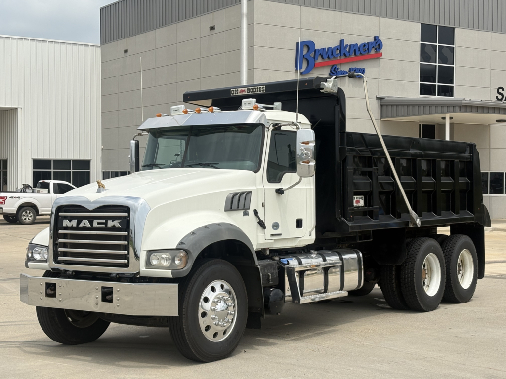 Mack GR64F dump truck in a dealership lot with white cab and black dump bed; visible text includes MACK on grille and Bruckner dealership building in background.