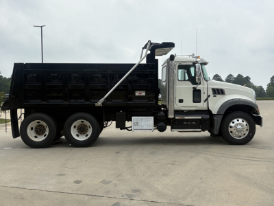 Dump truck side view showing 2020 Mack GR64F with dump bed, white cab, black dump body, chrome exhaust stack, dual rear axles, and fuel/tank area.