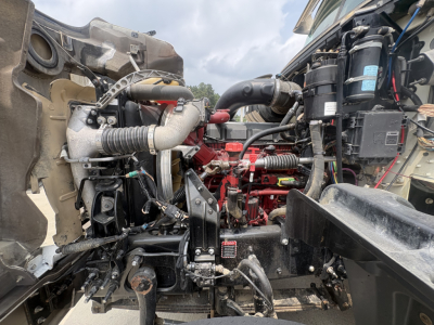 Mack GR64F engine bay showing diesel engine with red components, hoses, wiring, and surrounding chassis. Visible labels and hoses detail engine bay components.