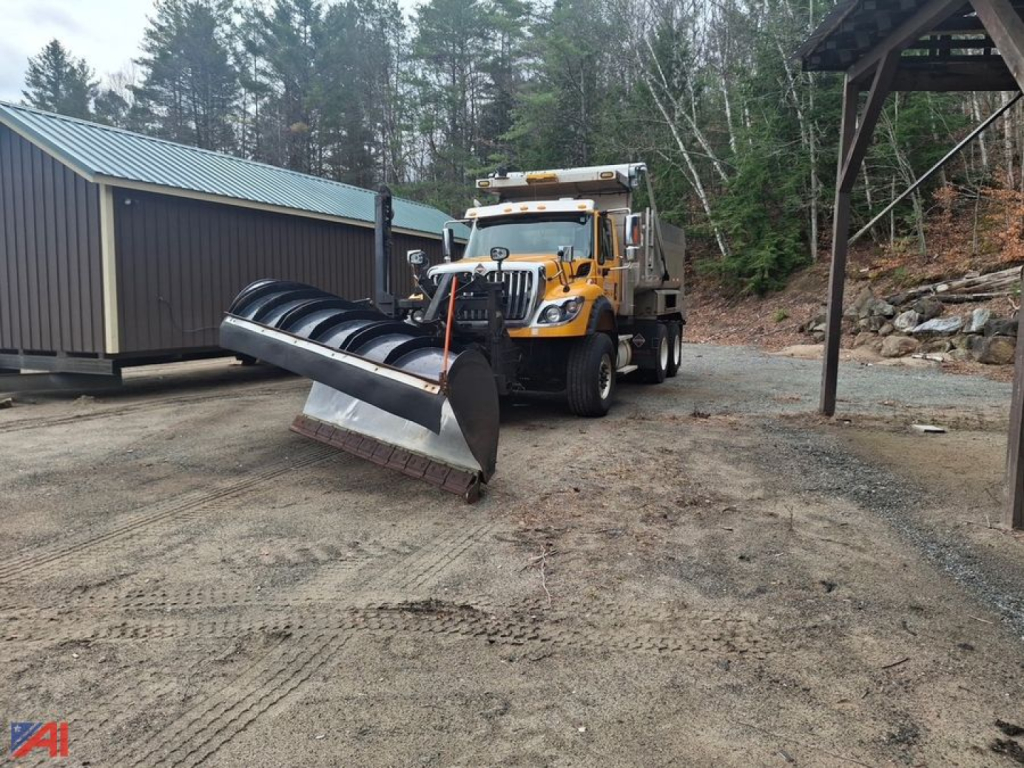 Front view of a 2017 International WorkStar 7600 dump truck with a large snowplow blade attached in the front, parked on gravel with a metal building and trees in the background.