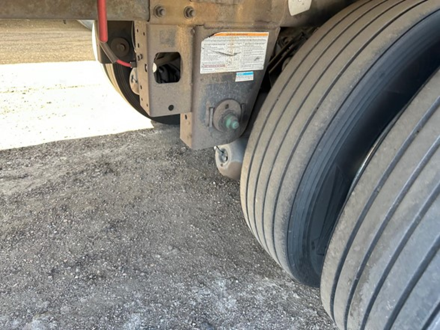 Trailer rear with dual tires and hydraulic components. Visible labels on frame with warning sticker and green component, plus wheel assembly.