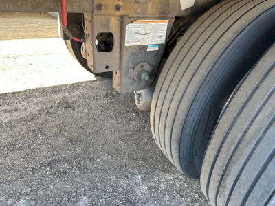 Trailer rear with dual tires and hydraulic components. Visible labels on frame with warning sticker and green component, plus wheel assembly.