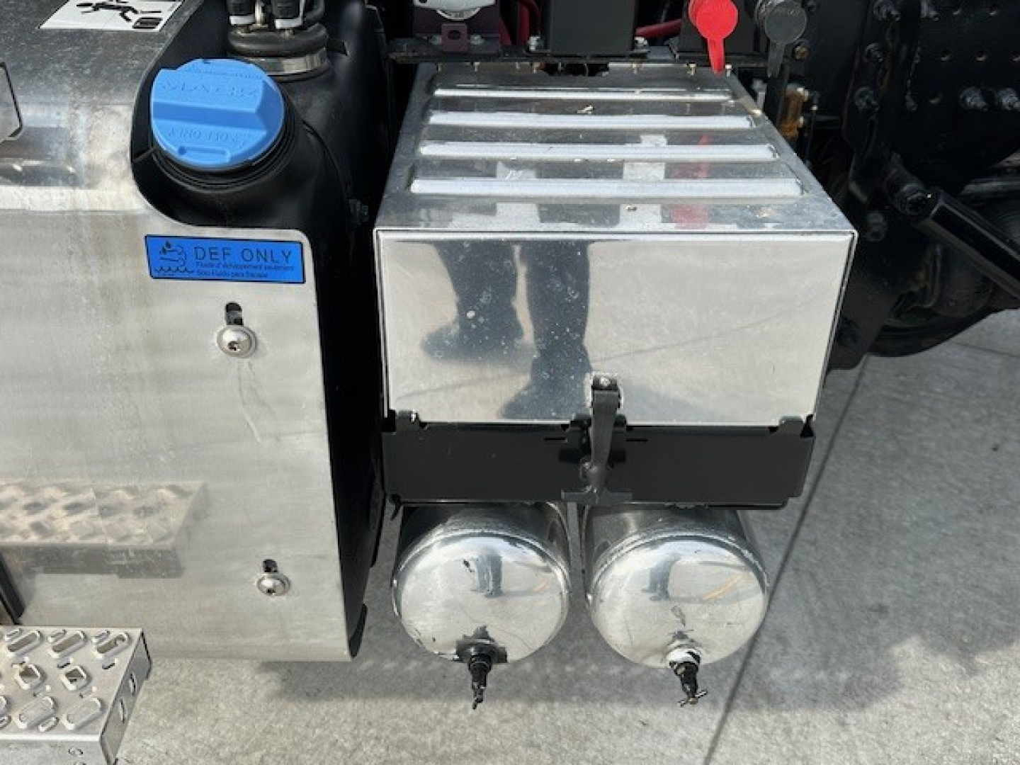 Truck component bay showing two round chrome fuel or sensor caps on a metal box with a blue cap reservoir labeled DEF ONLY and other metal toolbox, part of a Mack GR104F engine area.