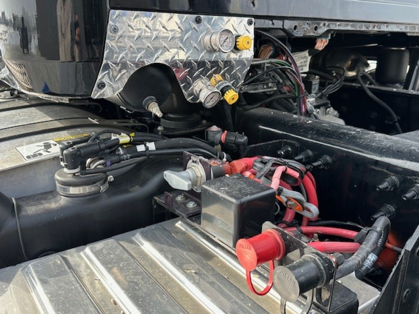 Truck engine bay showing battery terminals and wiring harnesses, access panel, and metal diamond plate with yellow caps in a Mack GR104F diesel truck.