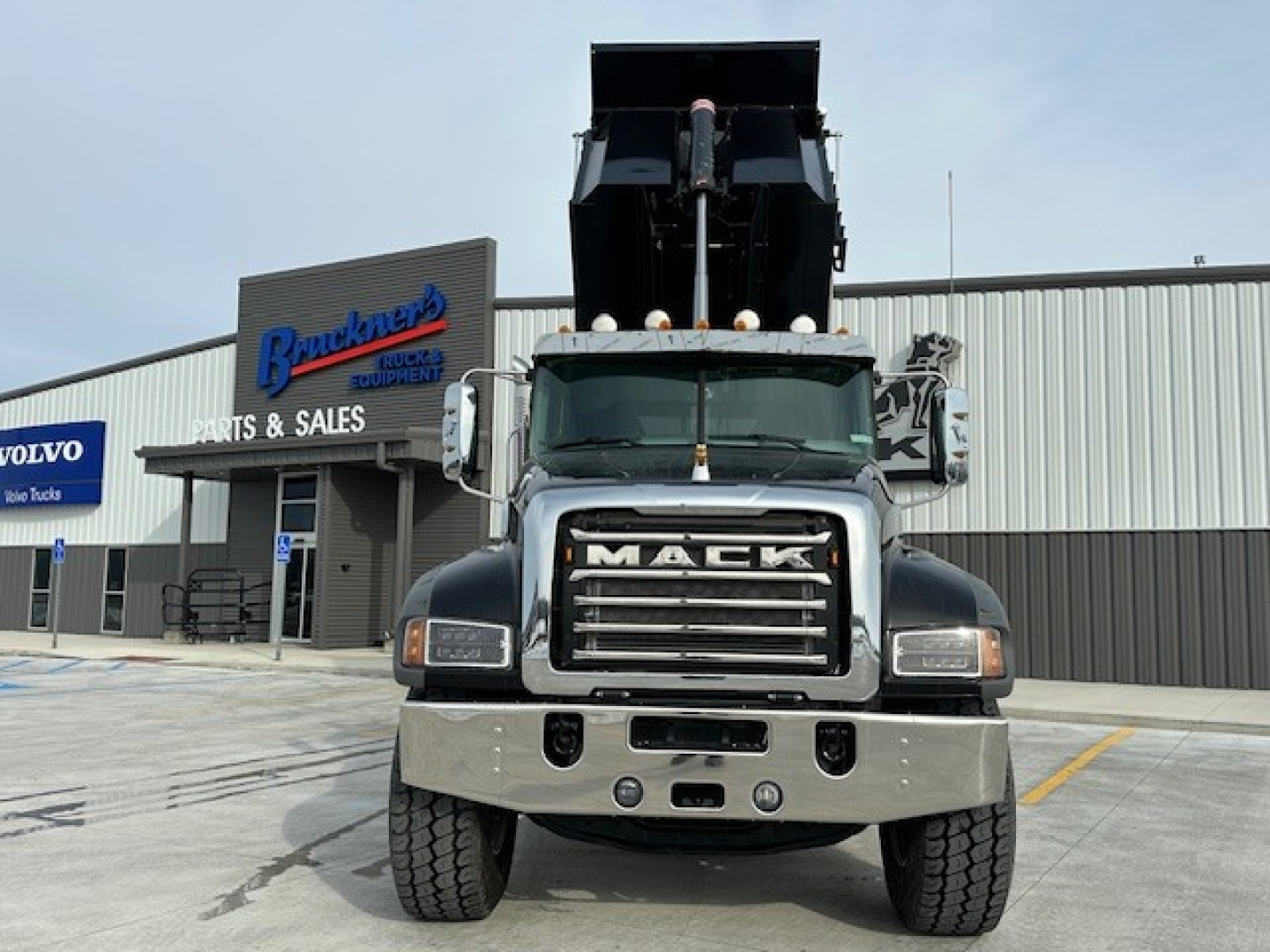 Dump truck front view showing Mack grille, headlights, chrome bumper, and raised dump bed in a dealership lot.