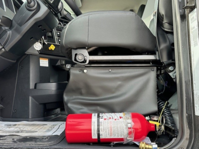 Cab interior of a Mack GR104F dump truck showing a red fire extinguisher mounted beneath the seat with protective cover and dashboard controls nearby.