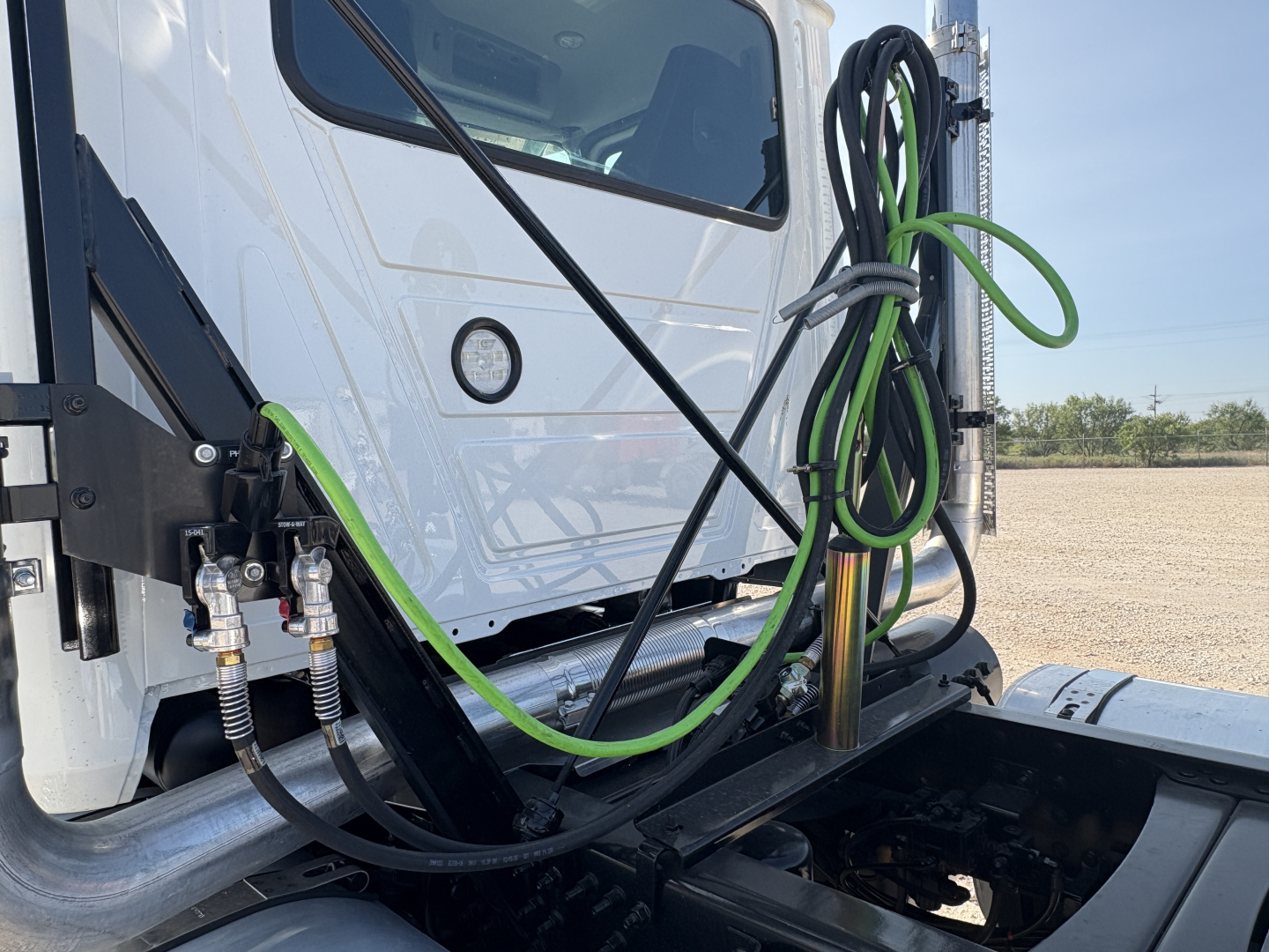 Front cab area of a 2027 Mack GR64FT showing green and black air lines, hydraulic hoses, and a chrome exhaust stack near the entry step; white tractor with window and VIN label visible.