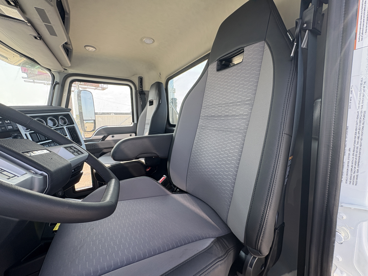 Truck cab interior showing Mack GR64FT seats and steering column, dashboard gauges, driver seat with gray fabric and black trim.