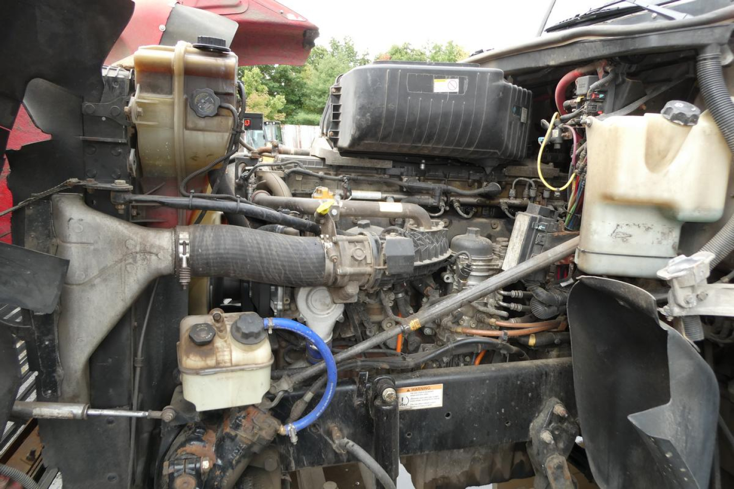Truck engine bay with various hoses, reservoirs, and components visible in a Freightliner SD122 cab area. Includes coolant reservoir, power steering lines, and air intake components.