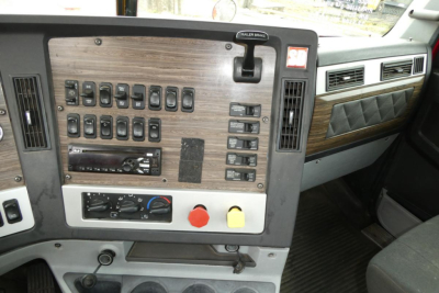 Dashboard panel of Freightliner SD122 cab with multiple switches, gauges, and control knobs; woodgrain panel and vent openings visible.
