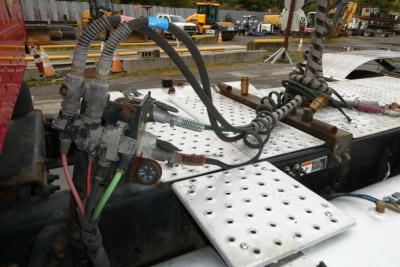 Conventional daycab engine bay area showing hydraulic lines, battery cables, and perforated metal step/deck surface with hoses and electrical connectors.
