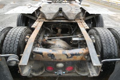 Rear view of a Freightliner SD122 chassis showing drivetrain, axles, and coupling—rusty frame with dual rear axles and fifth wheel area.